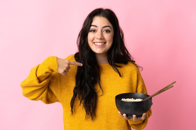 Joven mujer caucásica aislada en la pared rosa con expresión facial sorpresa mientras sostiene un plato de fideos con palillos