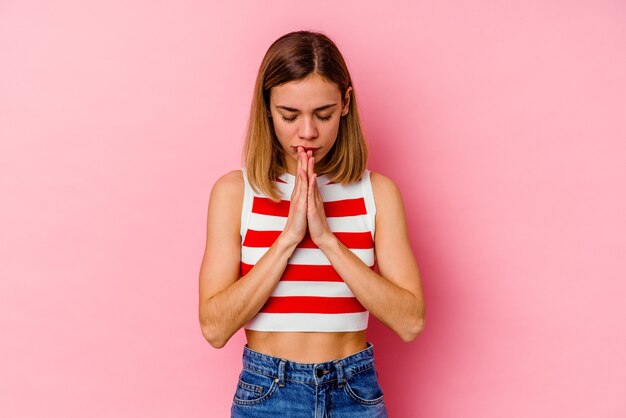 Joven mujer caucásica aislada en la pared rosa rezando, mostrando devoción, persona religiosa en busca de inspiración divina