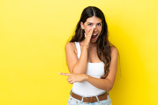 Joven mujer caucásica aislada sobre fondo amarillo apuntando hacia el lado para presentar un producto y susurrando algo