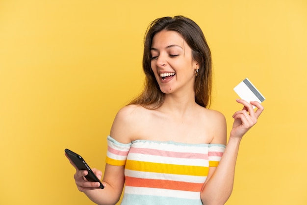 Joven mujer caucásica aislada sobre fondo amarillo comprando con el móvil con tarjeta de crédito