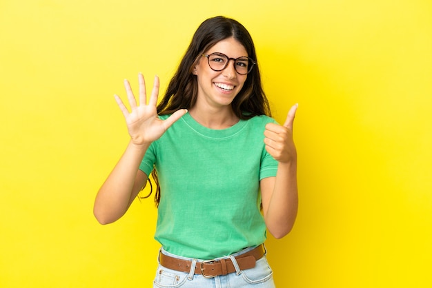 Joven mujer caucásica aislada sobre fondo amarillo contando seis con los dedos