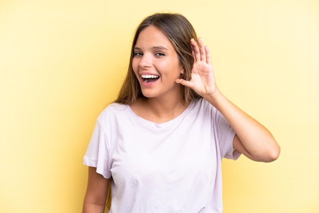 Joven mujer caucásica aislada sobre fondo amarillo escuchando algo poniendo la mano en la oreja