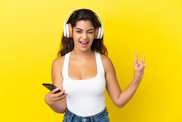 Joven mujer caucásica aislada sobre fondo amarillo escuchando música con un móvil haciendo gesto de rock