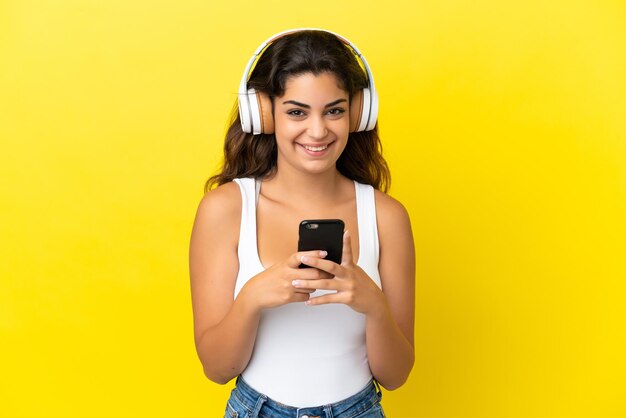 Joven mujer caucásica aislada sobre fondo amarillo escuchando música con un móvil y mirando al frente
