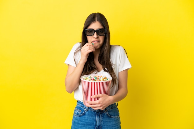 Joven mujer caucásica aislada sobre fondo amarillo con gafas 3d y sosteniendo un gran balde de palomitas de maíz