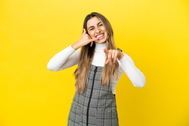 Joven mujer caucásica aislada sobre fondo amarillo haciendo gesto de teléfono y apuntando hacia el frente