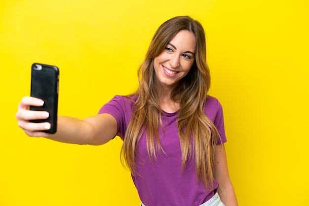 Joven mujer caucásica aislada sobre fondo amarillo haciendo un selfie con teléfono móvil