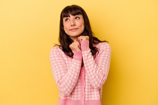 Joven mujer caucásica aislada sobre fondo amarillo mantiene las manos debajo de la barbilla, está mirando felizmente a un lado.