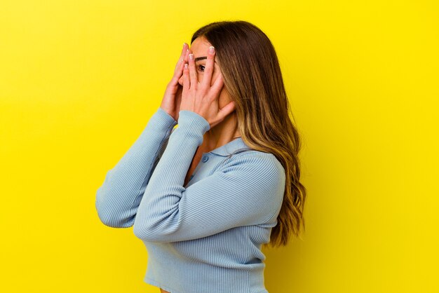 Joven mujer caucásica aislada sobre fondo amarillo parpadea a través de los dedos asustada y nerviosa.