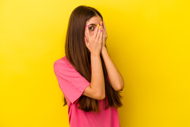 Joven mujer caucásica aislada sobre fondo amarillo parpadea a través de los dedos asustada y nerviosa.