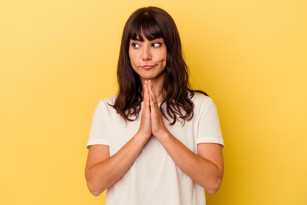 Joven mujer caucásica aislada sobre fondo amarillo rezando, mostrando devoción, persona religiosa en busca de inspiración divina.