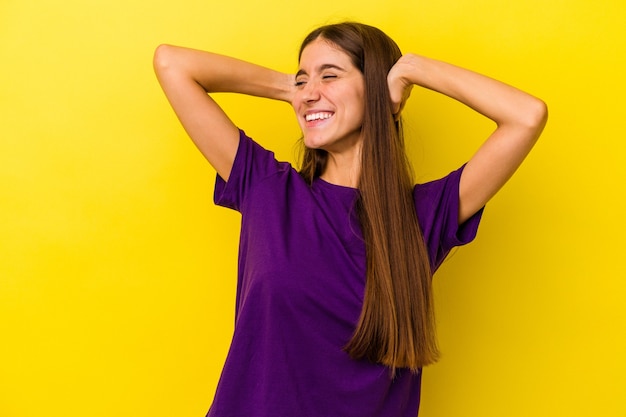 Joven mujer caucásica aislada sobre fondo amarillo sintiéndose confiada, con las manos detrás de la cabeza.