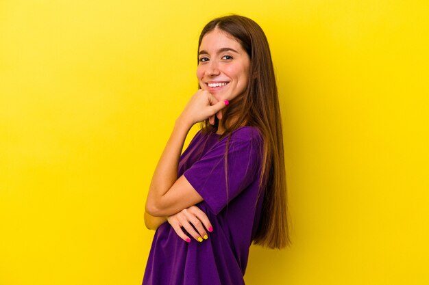 Joven mujer caucásica aislada sobre fondo amarillo sonriendo feliz y seguro, tocando la barbilla con la mano.