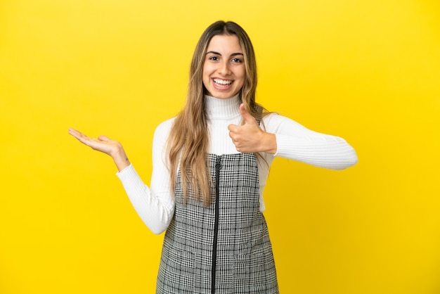 Joven mujer caucásica aislada sobre fondo amarillo sosteniendo copyspace imaginario en la palma para insertar un anuncio y con los pulgares hacia arriba