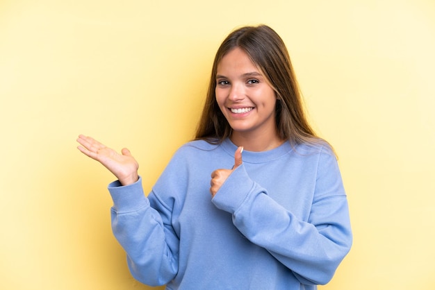 Joven mujer caucásica aislada sobre fondo amarillo sosteniendo copyspace imaginario en la palma para insertar un anuncio y con los pulgares hacia arriba