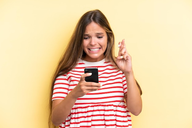 Joven mujer caucásica aislada sobre fondo amarillo mediante teléfono móvil con los dedos cruzando