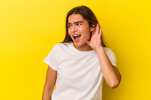 Joven mujer caucásica aislada sobre fondo amarillo tratando de escuchar un chisme.