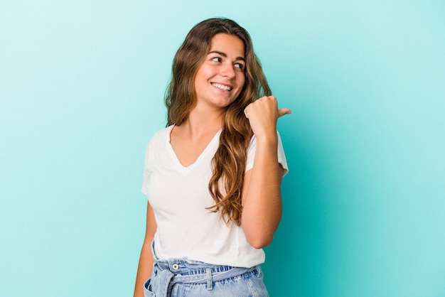 Joven mujer caucásica aislada sobre fondo azul apunta con el dedo pulgar lejos, riendo y despreocupado.