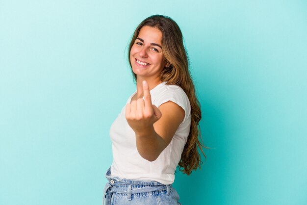 Joven mujer caucásica aislada sobre fondo azul apuntando con el dedo como si invitara a acercarse.