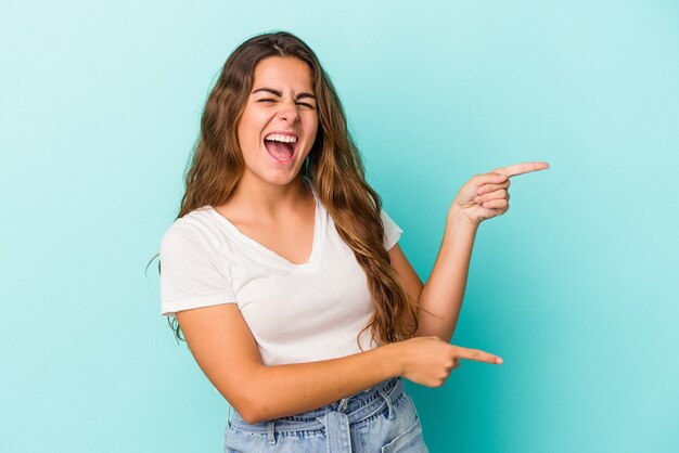 Joven mujer caucásica aislada sobre fondo azul apuntando con los dedos índices a un espacio de copia, expresando entusiasmo y deseo.