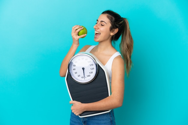 Joven mujer caucásica aislada sobre fondo azul con báscula y con una manzana