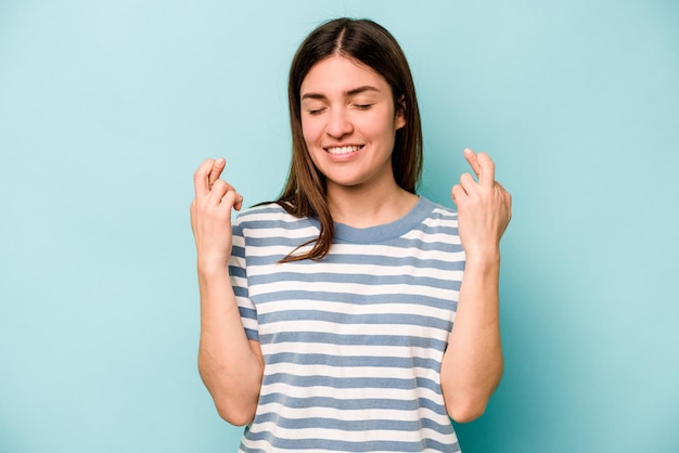 Joven mujer caucásica aislada sobre fondo azul cruzando los dedos para tener suerte