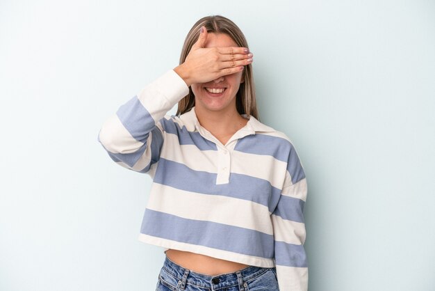 Joven mujer caucásica aislada sobre fondo azul cubre los ojos con las manos, sonríe ampliamente esperando una sorpresa.