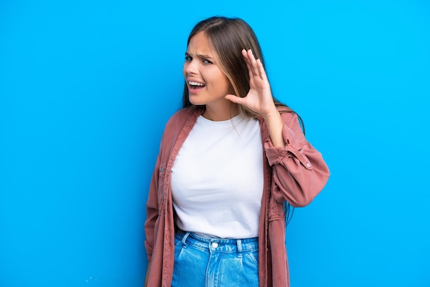 Joven mujer caucásica aislada sobre fondo azul escuchando algo poniendo la mano en la oreja