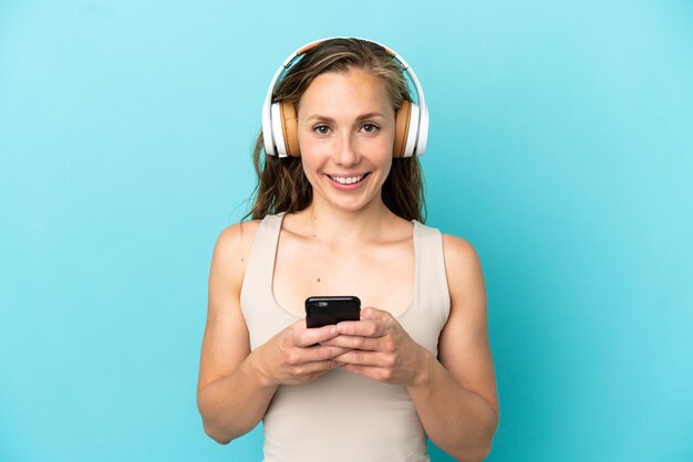 Joven mujer caucásica aislada sobre fondo azul escuchando música con un móvil y mirando al frente