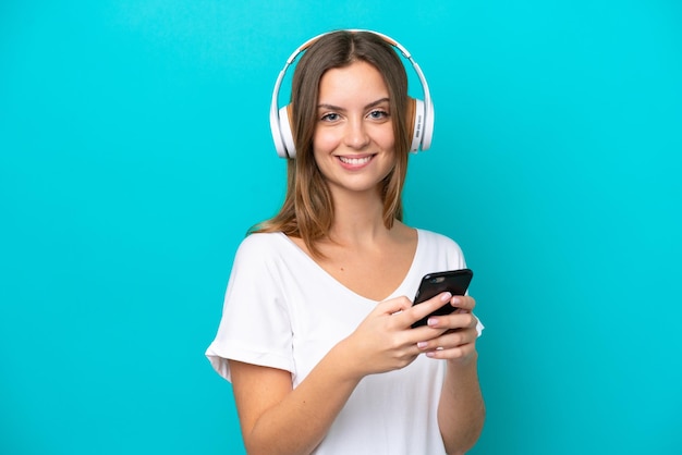 Joven mujer caucásica aislada sobre fondo azul escuchando música con un móvil y mirando al frente