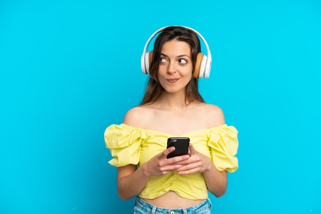 Joven mujer caucásica aislada sobre fondo azul escuchando música con un móvil y pensando