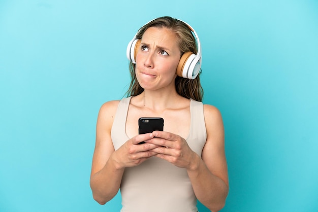Joven mujer caucásica aislada sobre fondo azul escuchando música con un móvil y pensando