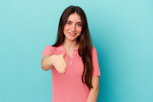 Joven mujer caucásica aislada sobre fondo azul estirando la mano a la cámara en gesto de saludo.