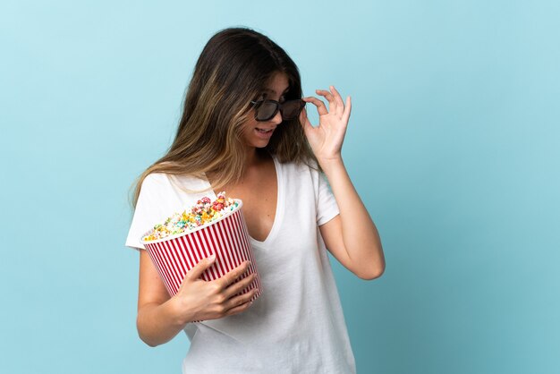 Joven mujer caucásica aislada sobre fondo azul con gafas 3d y sosteniendo un gran balde de palomitas de maíz