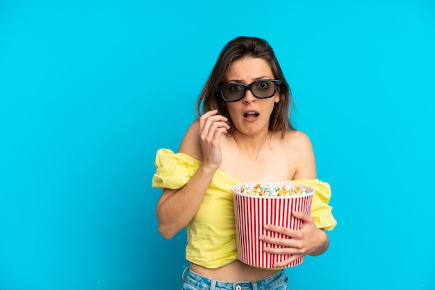 Joven mujer caucásica aislada sobre fondo azul con gafas 3d y sosteniendo un gran balde de palomitas de maíz