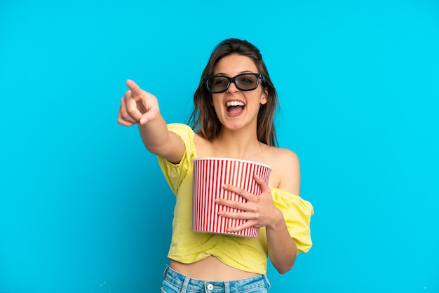 Joven mujer caucásica aislada sobre fondo azul con gafas 3d y sosteniendo un gran cubo de palomitas de maíz mientras apunta lejos