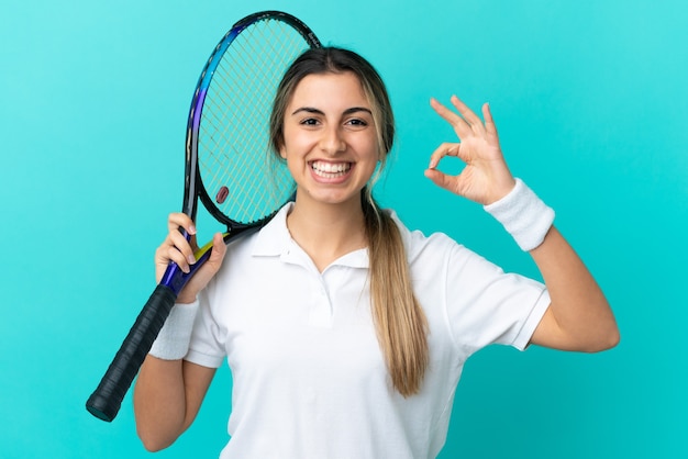 Foto joven mujer caucásica aislada sobre fondo azul jugando al tenis y haciendo el signo de ok