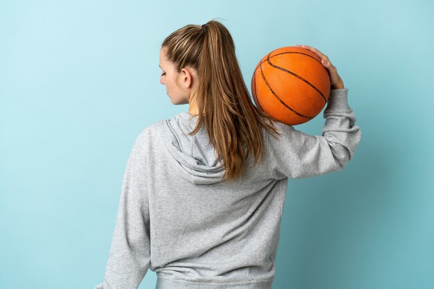 Joven mujer caucásica aislada sobre fondo azul jugando baloncesto en posición trasera