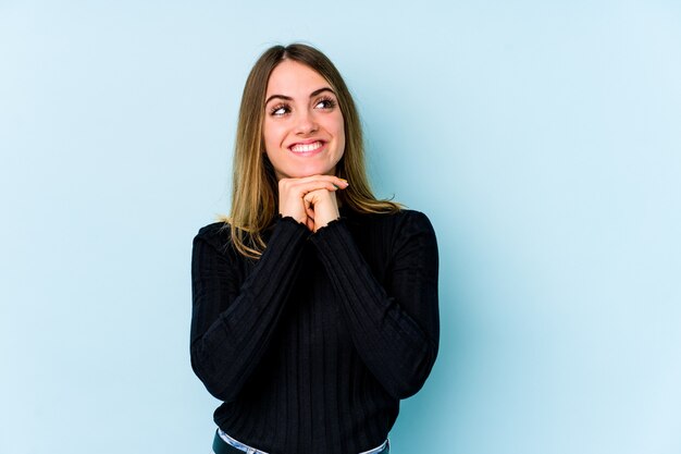 Joven mujer caucásica aislada sobre fondo azul mantiene las manos debajo de la barbilla, está mirando felizmente a un lado.