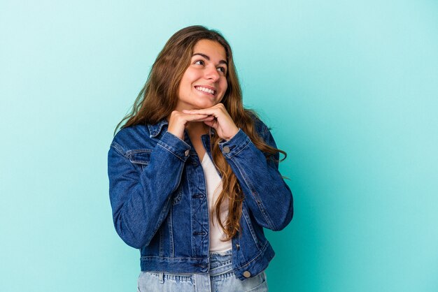 Joven mujer caucásica aislada sobre fondo azul mantiene las manos debajo de la barbilla, está mirando felizmente a un lado.