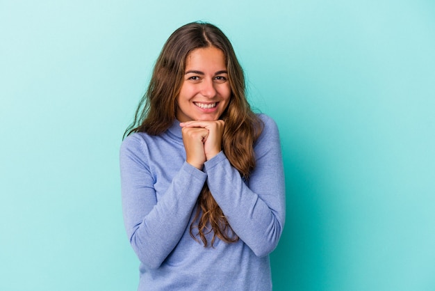 Joven mujer caucásica aislada sobre fondo azul mantiene las manos debajo de la barbilla, está mirando felizmente a un lado.