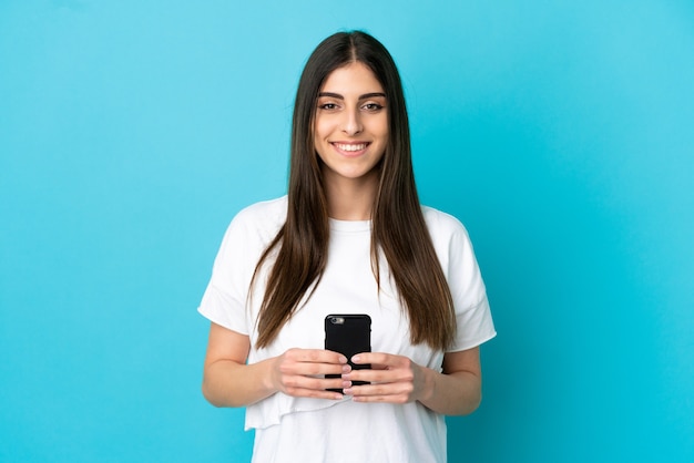 Foto joven mujer caucásica aislada sobre fondo azul mirando a la cámara y sonriendo mientras usa el móvil