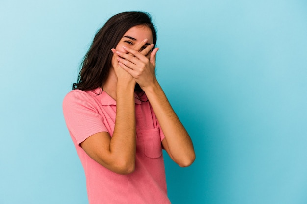 Joven mujer caucásica aislada sobre fondo azul parpadea a la cámara a través de los dedos, avergonzado cubriendo la cara.