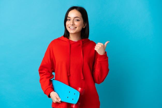 Joven mujer caucásica aislada sobre fondo azul con un patín y apuntando hacia el lado