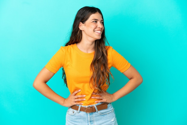 Foto joven mujer caucásica aislada sobre fondo azul posando con los brazos en la cadera y sonriendo