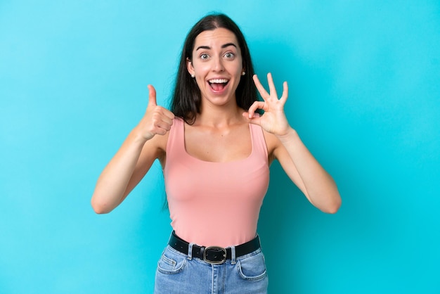 Joven mujer caucásica aislada sobre fondo azul que muestra el signo de ok y el pulgar hacia arriba gesto