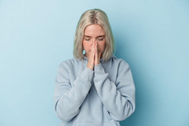 Joven mujer caucásica aislada sobre fondo azul rezando, mostrando devoción, persona religiosa en busca de inspiración divina.