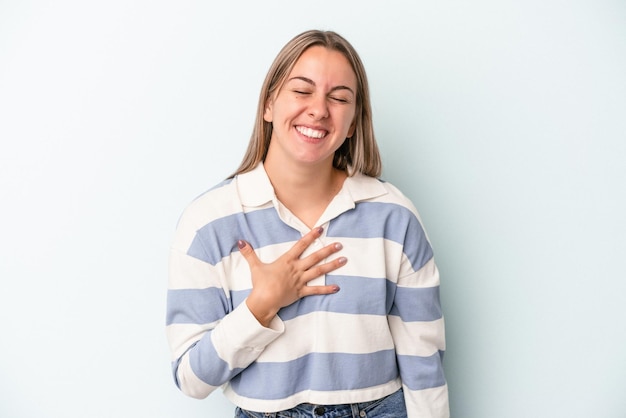 Joven mujer caucásica aislada sobre fondo azul se ríe a carcajadas manteniendo la mano en el pecho.