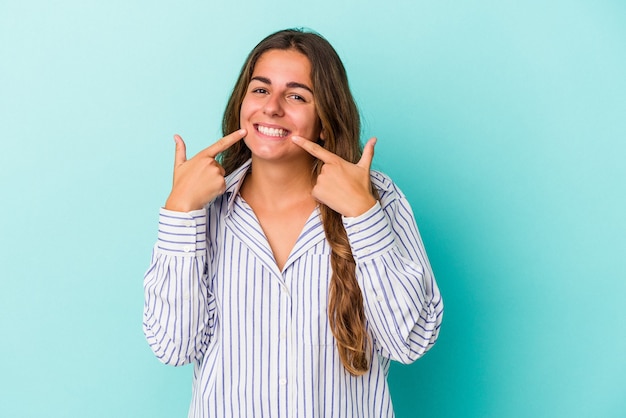 Joven mujer caucásica aislada sobre fondo azul sonríe, señalando con el dedo a la boca.