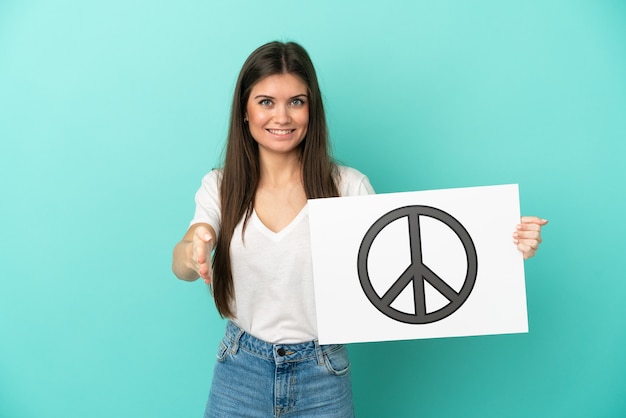 Joven mujer caucásica aislada sobre fondo azul sosteniendo un cartel con el símbolo de la paz haciendo un trato